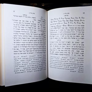 May include: An open book with Hebrew script, displaying black text on white pages. The book has a dark blue spine, and the pages are slightly off-white. The background is black, emphasizing the book's details.