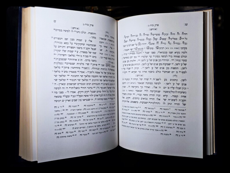 May include: An open book with Hebrew script, displaying black text on white pages. The book has a dark blue spine, and the pages are slightly off-white. The background is black, emphasizing the book's details.