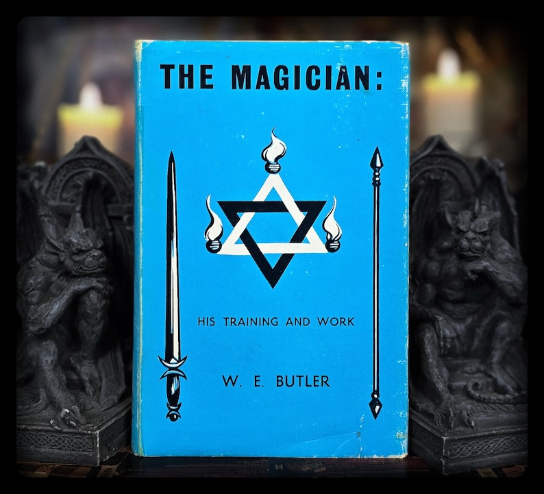 May include: A vintage book titled "THE MAGICIAN: HIS TRAINING AND WORK" by W. E. Butler. The book cover is light blue with a black and white Star of David design, flames, a sword, and a staff. The book is flanked by two dark gargoyle statues.