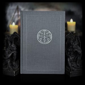 May include: A dark grey hardcover book featuring a silver circular seal on the front cover. The seal contains intricate symbols and the text "URIEL", "ADONAY", "ELOHIM", and "AGLA". The book is positioned between two dark stone gargoyle statues.