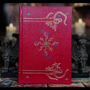 Puede incluir: Un libro encuadernado en cuero rojo con diseños decorativos dorados y azules. La portada presenta un árbol estilizado con una estrella en el centro.