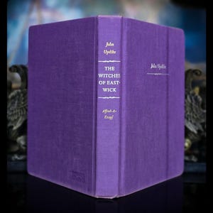 May include: A purple hardcover book titled The Witches of Eastwick by John Updike. The spine features gold lettering with the author name, title, and publisher Alfred A. Knopf. The front cover also displays the author name in gold script text.