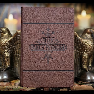 May include: A vintage brown book titled "OUR FAMILY PHYSICIAN" with decorative black text and embellishments. The book is upright, with a textured cover. Two golden eagle statues flank the book, with lit candles in the background.