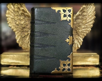May include: A dark green leather-bound book with gold-colored corner accents and a clasp. The book is set against a backdrop of golden angel wings and a golden base, creating a vintage aesthetic.