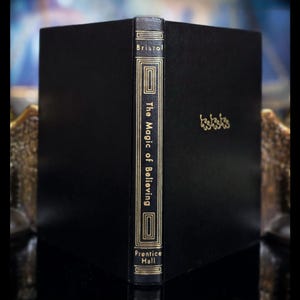 May include: A black hardcover book titled The Magic of Believing by Bristol, published by Prentice Hall. The spine features gold lettering and geometric border details, while the front cover displays three small gold oak leaf icons.