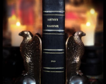 Può includere: Un libro rilegato in pelle nera con dettagli dorati e il titolo "SARTAIN'S MAGAZINE" e l'anno "1849" stampati sul dorso. Il libro è appoggiato su un paio di fermacarte a forma di aquila.