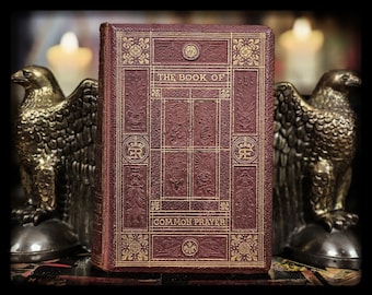 May include: A maroon-colored book titled "THE BOOK OF COMMON PRAYER" with gold embossed lettering and decorative patterns. The book is flanked by two gold eagle statues. The book's cover features intricate designs and a regal emblem.