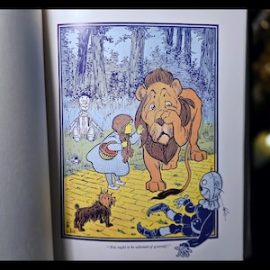 Puede incluir: Una colorida ilustraci&oacute;n de un libro infantil representa una escena de El Mago de Oz. Dorothy, con un vestido azul, interact&uacute;a con un gran le&oacute;n. Otros personajes incluyen un espantap&aacute;jaros, un hombre de hojalata y un perro peque&ntilde;o. El fondo muestra un bosque y un camino de baldosas amarillas.