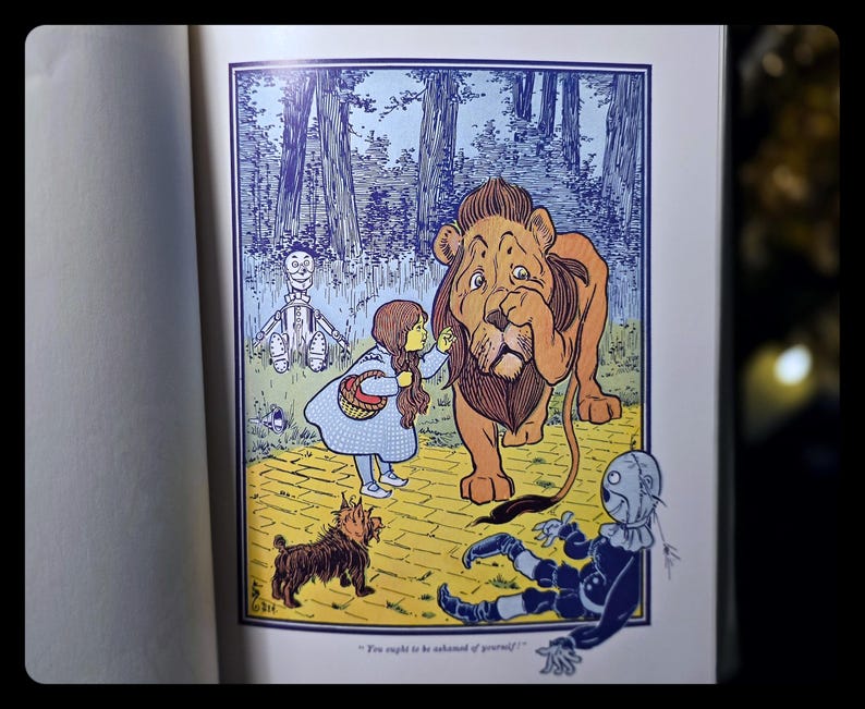 Puede incluir: Una colorida ilustraci&oacute;n de un libro infantil representa una escena de El Mago de Oz. Dorothy, con un vestido azul, interact&uacute;a con un gran le&oacute;n. Otros personajes incluyen un espantap&aacute;jaros, un hombre de hojalata y un perro peque&ntilde;o. El fondo muestra un bosque y un camino de baldosas amarillas.