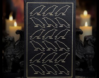 Può includere: Un libro rilegato in pelle nera con un motivo a corvi in rilievo dorato. I corvi sono disposti in un motivo ripetitivo, creando un design simmetrico.
