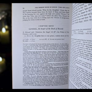 May include: An open book displaying text from "The Hebrew Book of Enoch". The page shows the chapter title "Chapter XXVII" and the name "Radweriel". The book is open on a dark surface, with a blurred background.