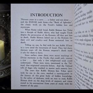 K&ouml;nnte beinhalten: Offenes Buch mit dem Titel "INTRODUCTION" und Text &uuml;ber den Zohar und Rabbi Shimon ben Yohai. Eine brennende Kerze in einem dekorativen Halter befindet sich im Vordergrund und erzeugt eine warme, spirituelle Atmosph&auml;re.