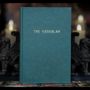 K&ouml;nnte beinhalten: Ein teal-farbenes Buch mit dem goldenen Titel "THE KABBALAH". Das Buch hat einen strukturierten Einband und steht aufrecht. Im Hintergrund befinden sich zwei steinerne Greifenstatuen und verschwommene Kerzen, was eine mystische Atmosph&auml;re erzeugt.