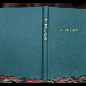 K&ouml;nnte beinhalten: Ein aufgeschlagenes Buch mit t&uuml;rkisfarbenem Einband, betitelt "THE KABBALAH" in goldener Schrift. Der Titel ist auch vertikal auf dem Buchr&uuml;cken aufgedruckt. Der Einband des Buches hat eine strukturierte Oberfl&auml;che. Der Hintergrund ist dunkel.