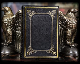 May include: A black book with gold trim and decorative accents stands between two bronze eagle bookends. The book has a rectangular gold-trimmed frame on the cover. The eagle bookends have detailed feather and wing designs.