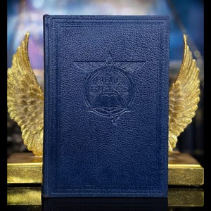 May include: A dark blue hardcover book with a textured leather-like finish. The front cover features an embossed circular emblem with the text The Bible of Bibles, centered beneath a winged symbol and above an open book illustration.