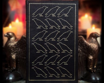 Può includere: Un libro rilegato in pelle nera con un motivo di corvi ripetuti in rilievo dorato. Il libro è su una superficie di legno con due statue di aquile decorative su entrambi i lati.