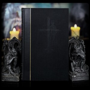 May include: A black hardcover book featuring a gold vertical line on the spine edge and a subtle embossed line drawing of a figure in a top hat standing before a cross on the front cover. The book is positioned upright with a dark, textured finish.