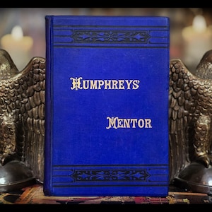 May include: A vintage book with a vibrant blue cover, titled "Humphreys Mentor" in gold lettering. Flanking the book are two bronze eagle statues. The book's cover features decorative black accents.