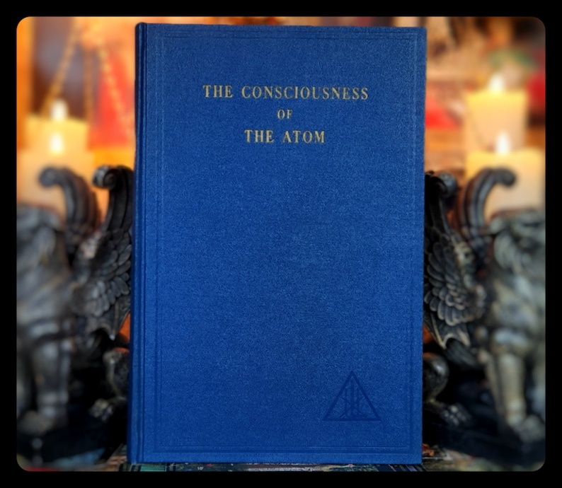 May include: A blue hardback book with the title "THE CONSCIOUSNESS OF THE ATOM" in gold lettering. A gold triangle symbol is embossed on the bottom of the cover.