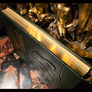 Puede incluir: Un libro verde oscuro con bordes dorados y detalles en relieve en la cubierta. El libro est&aacute; parcialmente abierto, revelando las p&aacute;ginas doradas. El fondo incluye una estatua dorada y una baraja de cartas del tarot, lo que sugiere un tema m&iacute;stico.