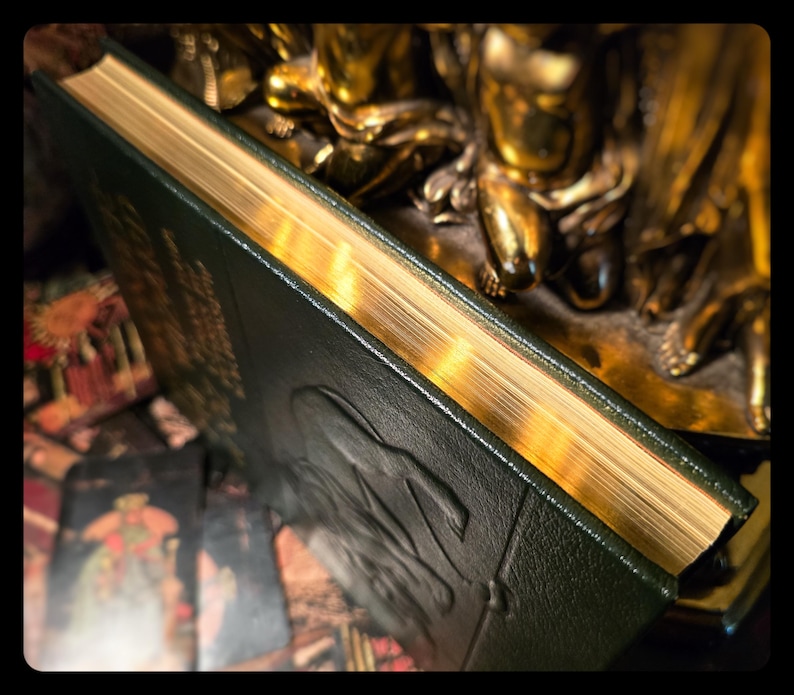 Puede incluir: Un libro verde oscuro con bordes dorados y detalles en relieve en la cubierta. El libro est&aacute; parcialmente abierto, revelando las p&aacute;ginas doradas. El fondo incluye una estatua dorada y una baraja de cartas del tarot, lo que sugiere un tema m&iacute;stico.