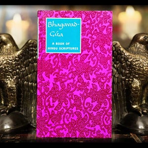 May include: A hardcover book titled "Bhagavad-Gita" with a bright pink floral pattern cover. The title is in a blue box. Two gold eagle statues flank the book, creating a symmetrical composition. The book is a book of Hindu scriptures.