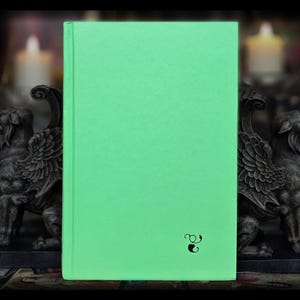 May include: A hardcover book with a bright green cover. A small black illustration is in the lower right corner. The book is set against a dark background with decorative statues and lit candles.