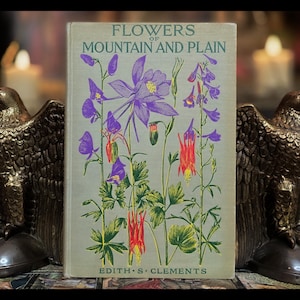 May include: A vintage book titled "Flowers of Mountain and Plain" by Edith S. Clements. The cover features a colorful illustration of various wildflowers, including purple columbine, red and white flowers, and green foliage.
