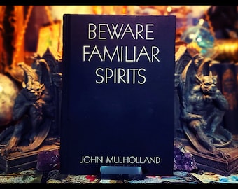 May include: A black book with the title "Beware Familiar Spirits" and the author's name "John Mulholland" printed in white on the front cover. The book is displayed on a stand with a dark purple crystal on either side.