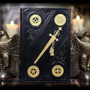 Könnte beinhalten: Ein schwarzes Buch mit goldgeprägten Freimaurersymbolen und einem Schwertdesign. Das Buch wird von zwei bronzenen Adlerstatuen flankiert, mit brennenden Kerzen im Hintergrund. Der Einband des Buches weist komplizierte Details und ein dekoratives Muster auf.