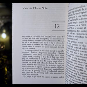 May include: Open book page with the title "Scientists Please Note" and the number "12" in bold. The text discusses scientific concepts and the nature of the universe. The book's pages are off-white.