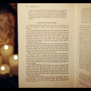 May include: Open book with a yellowed page, showing text about reincarnation and the duration of time between lives. The book is open to a page with the title "How Many Lifetimes?" and the text discusses the concept of souls returning to Earth after death. The book is illuminated by two candles, casting a warm glow on the page.
