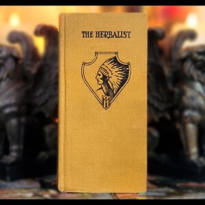 May include: A vintage book titled "The Herbalist" with a yellow linen cover and a black and white illustration of a Native American wearing a headdress on the front.