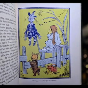Puede incluir: Ilustraci&oacute;n de libro vintage de El Mago de Oz. La imagen muestra a Dorothy, el Espantap&aacute;jaros, Toto y una cesta. La ilustraci&oacute;n es en tonos de azul, amarillo y marr&oacute;n, con texto en la p&aacute;gina izquierda.