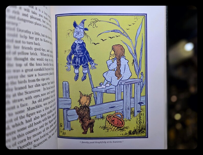 Puede incluir: Ilustraci&oacute;n de libro vintage de El Mago de Oz. La imagen muestra a Dorothy, el Espantap&aacute;jaros, Toto y una cesta. La ilustraci&oacute;n es en tonos de azul, amarillo y marr&oacute;n, con texto en la p&aacute;gina izquierda.