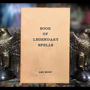 Puede incluir: Un libro de color beige con el título "Book of Legendary Spells" de Elbee Wright. El libro se muestra entre dos sujetalibros de águila dorada.