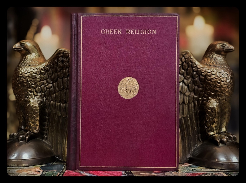 May include: A burgundy hardcover book with gold lettering that reads "Greek Religion". The book is sitting on a table with two golden eagle statues on either side.