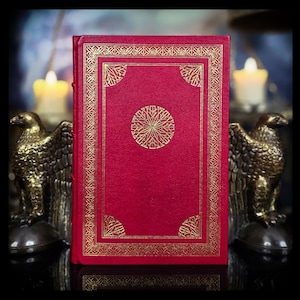 May include: A hardcover book with a vibrant red cover featuring intricate gold foil stamping. The design includes a rectangular border with corner accents and a central circular motif. The book is positioned between two bronze eagle statues.