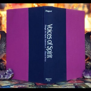 Puede incluir: Un libro de tapa dura de color morado y azul marino titulado "Voices of Spirit: Through the Psychic Experiences of Elwood Babbitt" de Hagood, publicado por DeVorss Press.