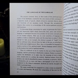 K&ouml;nnte beinhalten: Offenes Buch mit dem Titel "THE LANGUAGE OF THE KABBALAH" und einer brennenden Kerze im Hintergrund. Der Text behandelt die Verwendung von bildhafter Sprache und Symbolen in der Kabbala zur Vermittlung spiritueller Konzepte.