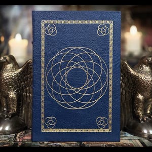 May include: A dark blue book with gold geometric designs and accents. The cover features a large circular pattern and smaller corner designs. The book is flanked by two bronze eagle statues, with lit candles in the background, creating a mystical aesthetic.