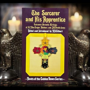 May include: A book titled "The Sorcerer and His Apprentice" with a purple cover and gold lettering. The book features a colorful cross-shaped pendant. Two bronze eagle statues flank the book, with lit candles in the background.