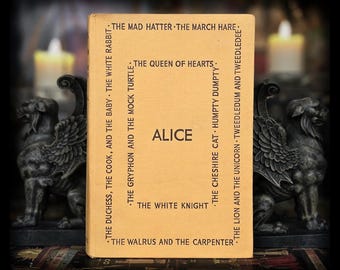 May include: A vintage book titled "Alice" with a light tan cover. The book's spine and cover feature text including character names like "The Mad Hatter" and "The Queen of Hearts." Two decorative griffin statues flank the book, with blurred candles in the background.
