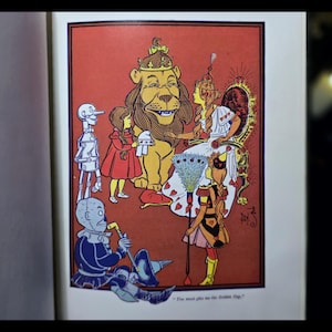 Puede incluir: Ilustraci&oacute;n vintage de un libro infantil con personajes de El Mago de Oz. La imagen muestra al Le&oacute;n Cobarde, Dorothy, el Hombre de Hojalata y el Espantap&aacute;jaros, sobre un fondo rojo.