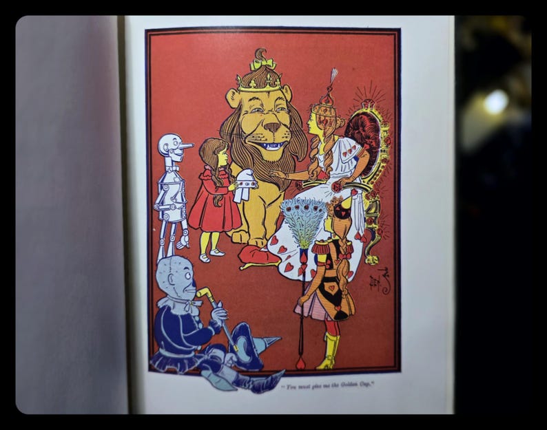 Puede incluir: Ilustraci&oacute;n vintage de un libro infantil con personajes de El Mago de Oz. La imagen muestra al Le&oacute;n Cobarde, Dorothy, el Hombre de Hojalata y el Espantap&aacute;jaros, sobre un fondo rojo.
