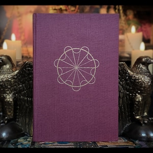 Pode incluir: Um livro de capa dura de cor roxo escuro com um design geom&eacute;trico dourado na capa. O design &eacute; um c&iacute;rculo com oito pontos e uma estrela central.