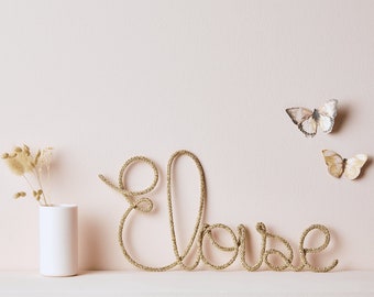 May include: A gold-colored wire word "Eloise" is displayed on a white shelf against a light pink wall. Two butterfly decorations are affixed to the wall. A small white vase holds dried flowers to the left of the word.