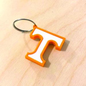 May include: Orange and white plastic keychain with a large letter "T" in a white outline. The keychain has a silver metal ring.