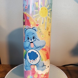 May include: A tall, insulated tumbler with a colorful design featuring a blue Care Bear sticking out its tongue. The tumbler has a rainbow, sun, and cloud design with a pink and yellow background.